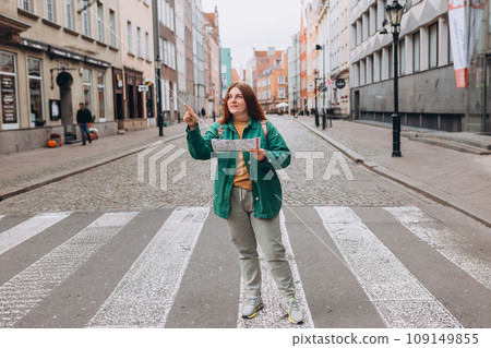 Casual dressed woman 30 years old exploring city streets during international excursion in town. Redhead girl with backpack pointing finger on city street. Traveling Europe in autumn. Full body Casual dressed woman 30 years old exploring city streets during international excursion in town. Redhead girl with backpack pointing finger on city street. Traveling Europe in autumn. Full body 109149855