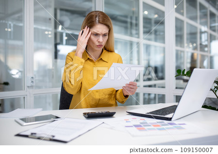Serious young business woman working in the office at the laptop, holding documents in her hands and holding her head worriedly. 109150007