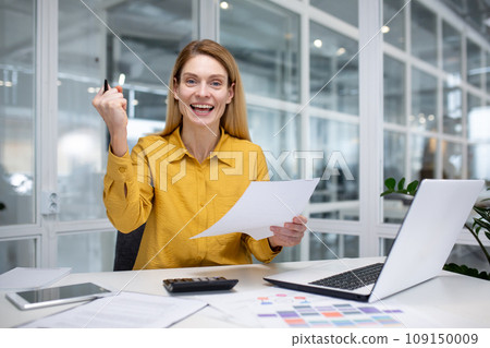 Portrait of a happy businesswoman working in the office at a table with a laptop, holding a document in her hands, showing a sign of success and smiling and looking at the camera. Portrait of a happy businesswoman working in the office at a table with a laptop, holding a document in her hands, showing a sign of success and smiling and looking at the camera. 109150009