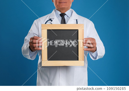 Doctor holding blackboard with word HEMORRHOID on blue background, closeup Doctor holding blackboard with word HEMORRHOID on blue background, closeup 109150285