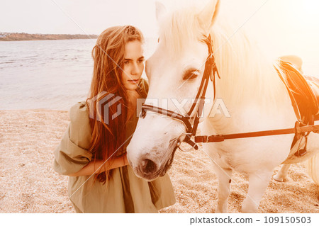 A woman in a dress stands next to a white horse on a beach, with the blue sky and sea in the background. A woman in a dress stands next to a white horse on a beach, with the blue sky and sea in the background. 109150503