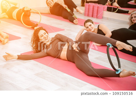 A group of six athletic women doing pilates or yoga on pink mats in front of a window in a beige loft studio interior. Teamwork, good mood and healthy lifestyle concept. A group of six athletic women doing pilates or yoga on pink mats in front of a window in a beige loft studio interior. Teamwork, good mood and healthy lifestyle concept. 109150516