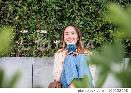 In a tranquil park on a lovely spring day a cute woman sits with her bicycle using her mobile phone to listen to music with earphones. Her joyful typing and bright smile capture essence of season. In a tranquil park on a lovely spring day a cute woman sits with her bicycle using her mobile phone to listen to music with earphones. Her joyful typing and bright smile capture essence of season. 109152498
