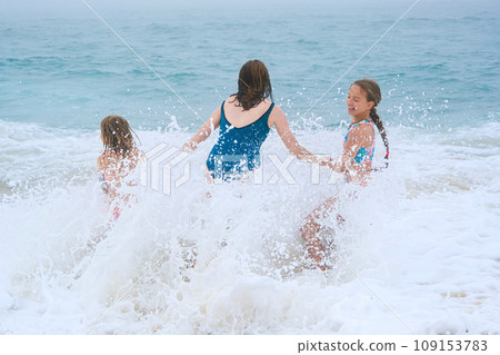 Mother and children playing on the ocean beach. Family enjoying the ocean. Mother holds girls's hands and they all look at the ocean together. Mother and children playing on the ocean beach. Family enjoying the ocean. Mother holds girls's hands and they all look at the ocean together. 109153783