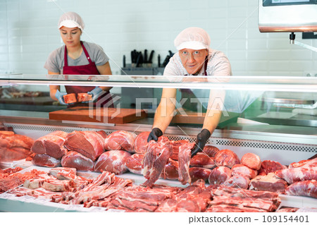 Elderly butcher shop seller laying out fresh beef in display case 109154401