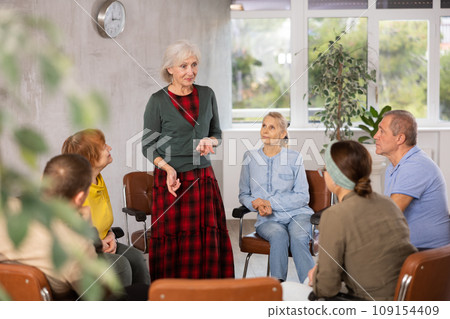 Older woman presides over support group meeting with mature people listening Older woman presides over support group meeting with mature people listening 109154409