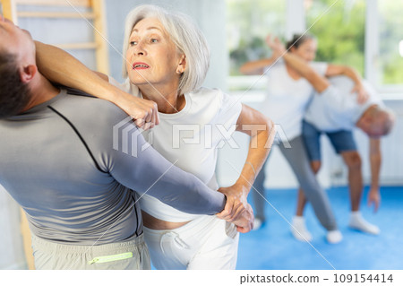 Elderly woman practices strikes and submissions against an attacking man during self-defense lesson in gym Elderly woman practices strikes and submissions against an attacking man during self-defense lesson in gym 109154414