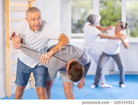 Senior man in pair with middle-aged coach train Krav Maga and self-defense skills. 109154581