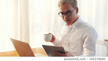 Asian Man drinking coffee cup using laptop at home office. Handsome man wear eyeglass holding smart tablet look website. Freelance Men using laptop home office technology. Businessman online meeting 109159680