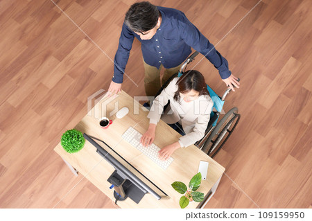 A woman in a wheelchair using a PC and a man supporting her A woman in a wheelchair using a PC and a man supporting her 109159950