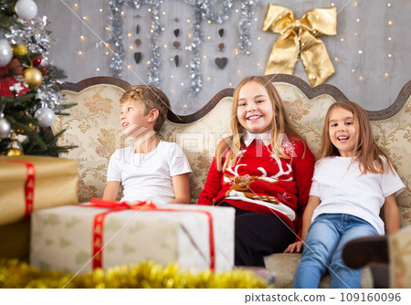 Happy brother with two sisters are sitting on sofa together with gift boxes next to the new year tree Happy brother with two sisters are sitting on sofa together with gift boxes next to the new year tree 109160096