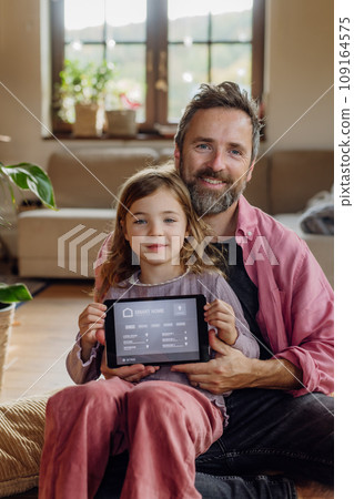 Father and girl holding smart thermostat, adjusting, lowering heating temperature at home. Concept of sustainable, efficient, and smart technology in home heating and thermostats. Father and girl holding smart thermostat, adjusting, lowering heating temperature at home. Concept of sustainable, efficient, and smart technology in home heating and thermostats. 109164575