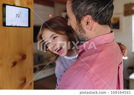 Father looking at smart thermostat, adjusting, lowering heating temperature at home. Concept of sustainable, efficient, and smart technology in home heating and thermostats. 109164581