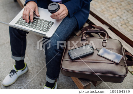 Businessman, freelancer or manager working outdoors in city park. Man with laptop on knees drinking coffee, charging his smartphone with solar phone charger. Concept of working remotely. 109164589