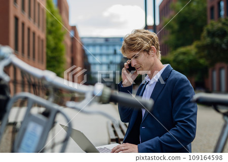 Businessman, freelancer or manager working outdoors in city park. Man with laptop on knees making call on smartphone, sitting on park bench during sunny day. Concept of working remotely. 109164598