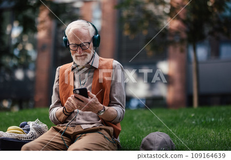 Stylish senior man sitting in the city park, listening to music via headphones. Concept of old man young at heart. 109164639