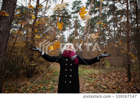Happy senior woman throws leaves in forest or natural park in autumn, smiles. Older female model stylishly dressed in black coat, scarf. Mature joy, active lifestyle Happy senior woman throws leaves in forest or natural park in autumn, smiles. Older female model stylishly dressed in black coat, scarf. Mature joy, active lifestyle 109167147