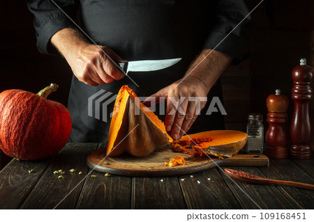 A chef cuts an orange pumpkin into slices with a knife on a wooden cutting board before preparing porridge for breakfast. Autumn food concept in cozy dark kitchen 109168451