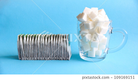 Many tea bags in a row next to a transparent glass mug full of white refined sugar on blue background Many tea bags in a row next to a transparent glass mug full of white refined sugar on blue background 109169151