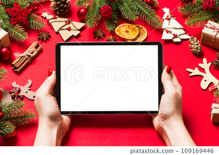 Top view of woman holding tablet in her hands on red background made of Christmas decorations. New Year holiday concept. Mockup Top view of woman holding tablet in her hands on red background made of Christmas decorations. New Year holiday concept. Mockup 109169446