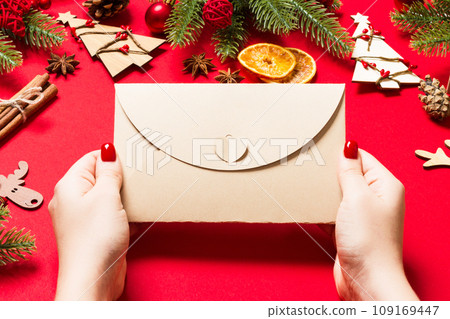 Top view of woman holding an envelope on red background made of holiday decorations. Christmas time concept Top view of woman holding an envelope on red background made of holiday decorations. Christmas time concept 109169447