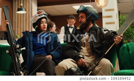 Travelers arrive in cozy ski resort lobby for skiing winter getaway waiting for room check in. Diverse people dressed in snow gear carrying ski or snowboard equipment at alpine hotel. Handheld shot. Travelers arrive in cozy ski resort lobby for skiing winter getaway waiting for room check in. Diverse people dressed in snow gear carrying ski or snowboard equipment at alpine hotel. Handheld shot. 109169452