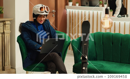 Tourist reading reviews on laptop at luxury hotel, sitting in lounge area with skiing equipment and accessories. Asian woman working remote on pc before practicing extreme winter sport in the cold. Tourist reading reviews on laptop at luxury hotel, sitting in lounge area with skiing equipment and accessories. Asian woman working remote on pc before practicing extreme winter sport in the cold. 109169458