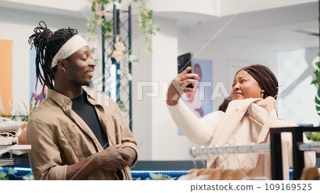 Man in fancy fashion boutique convincing his girlfriend to buy stylish formal clothes on hangers. Customers in premium showroom debating whether to purchase new designer clothing collection or not 109169525