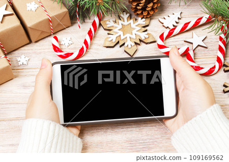 Christmas online shopping. Female buyer makes order at screen of smartphone with copy space. Woman buys presents for xmas eve, sits at home near decorated. Winter holidays sales 109169562