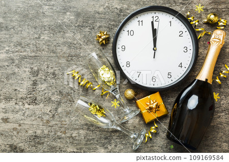 New year celebration concept with a bottle of champagne and two glasses toasting. Christmas gift box, alarm clock and fir tree branch on colored table. Top view Copy space 109169584