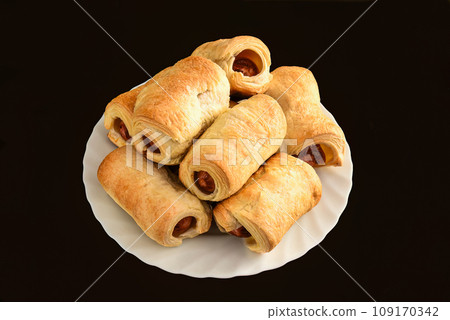 Many sausage rolls (pigs in a blanket) on white plate on black table 109170342