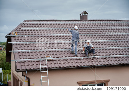 Men mounters prepearing for mounting photovoltaic solar moduls on roof of house. Electricians in helmets installing solar panel system outdoors. Concept of alternative and renewable energy. 109171000