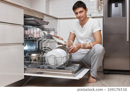 handsome positive man pulls out clean dishes from the dishwasher in white modern kitchen handsome positive man pulls out clean dishes from the dishwasher in white modern kitchen 109176538