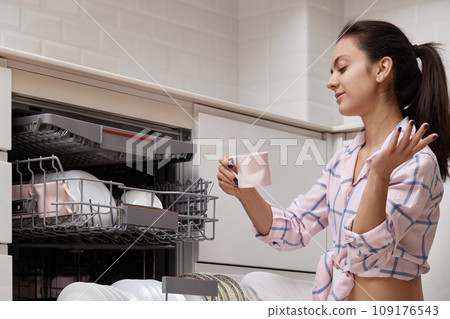 attractive woman unloading cup from open automatic built-in dishwasher machine with clean utensils inside in modern white kitchen. 109176543