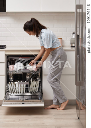 happy woman unloading plate from open automatic built-in dishwasher machine with clean utensils inside in modern white kitchen. happy woman unloading plate from open automatic built-in dishwasher machine with clean utensils inside in modern white kitchen. 109176548