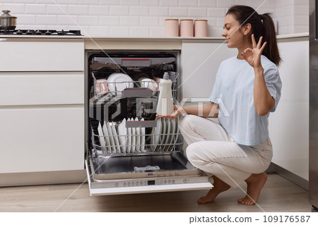 woman pours rinse aid into the dishwasher compartment in modern white kitchen 109176587