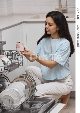 pretty woman unloading from open automatic built-in dishwasher machine with clean utensils inside in modern white kitchen. pretty woman unloading from open automatic built-in dishwasher machine with clean utensils inside in modern white kitchen. 109176609
