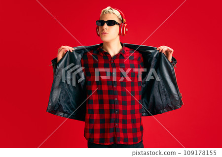 Stylish young man in checkered shirt, sunglasses and leather jacket standing and listening to music in headphones against red studio background 109178155