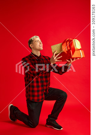 Young smiling happy guy in casual clothes standing on one knee and giving present box against red studio background. Christmas, winter holidays 109178163