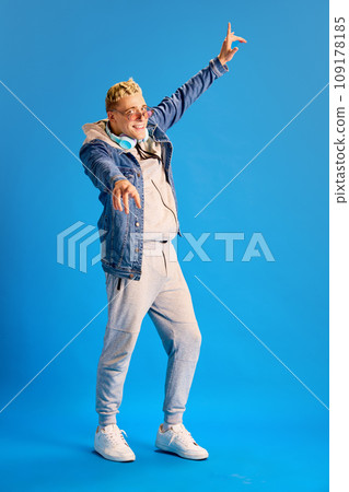 Young positive stylish guy in sunglasses, jeans jacket and casual sportswear, wit headphones, smiling against blue studio background 109178185
