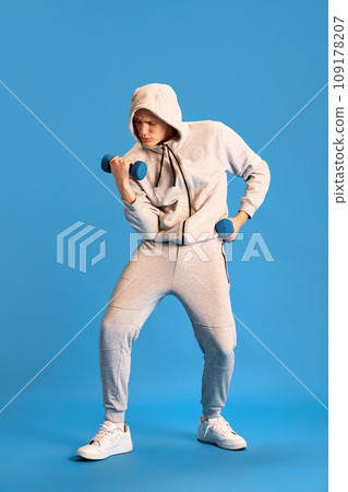 Strong hands. Young man in grey sportswear training, doing hand exercises with dumbbells against blue studio background 109178207