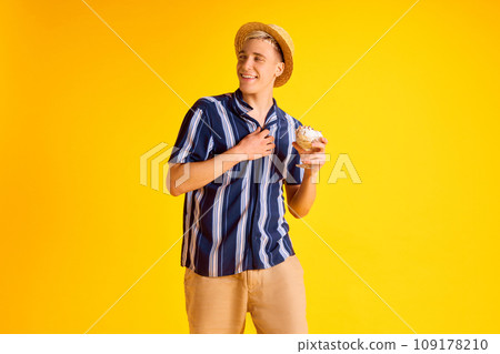 Handsome smiling young man in striped shirt, shorts and straw hat eating delicious vanilla ice cream against yellow studio background 109178210