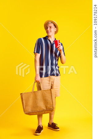 Stylish, young smiling man in shirt and shorts, holding bag, drinking coke against yellow studio background. Tourist 109178214