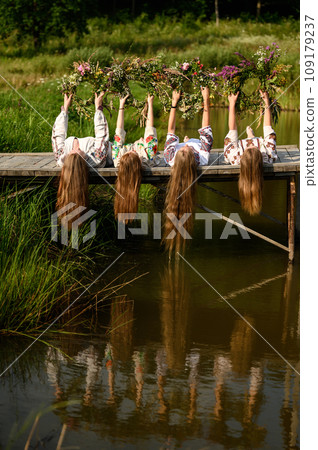 The girls are lying on the bridge near the pond and have raised woven wreaths above themselves. 109179237