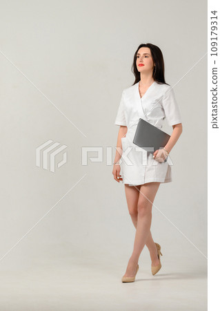 a cosmetologist girl in a white coat holds a laptop in her hands in a photo studio on a white background. 109179314