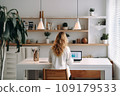 young woman working from home in front of a computer monitor. The apartment is minimalistic, with plants 109179533