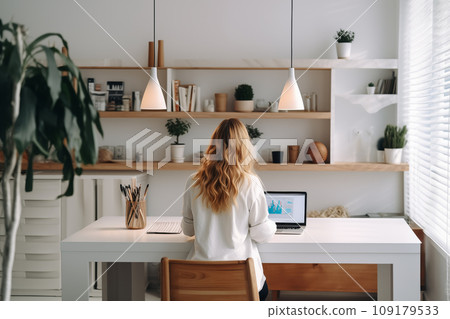 young woman working from home in front of a computer monitor. The apartment is minimalistic, with plants 109179533