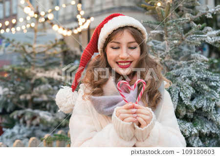 Young smiling woman in Santa hat making heart shape with candy canes while walking on traditional street Christmas market. Happy New Year celebration Christmas holidays. Share love, charity concept 109180603