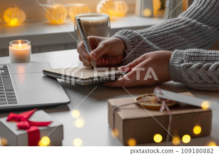 Young woman writing christmas cards, makes order on laptop. Female picking gifts online purchases at cozy home among gift boxes and packages. Winter sales, Black Friday. Christmas discount promotions Young woman writing christmas cards, makes order on laptop. Female picking gifts online purchases at cozy home among gift boxes and packages. Winter sales, Black Friday. Christmas discount promotions 109180882