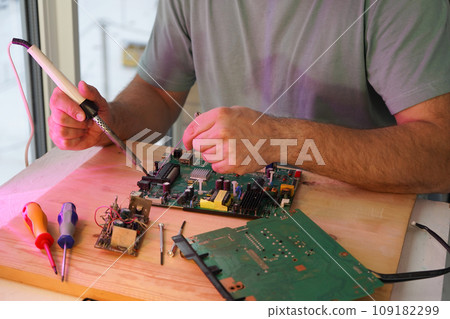 A man holds a soldering iron in his hand and solders a microcircuit A man holds a soldering iron in his hand and solders a microcircuit 109182299